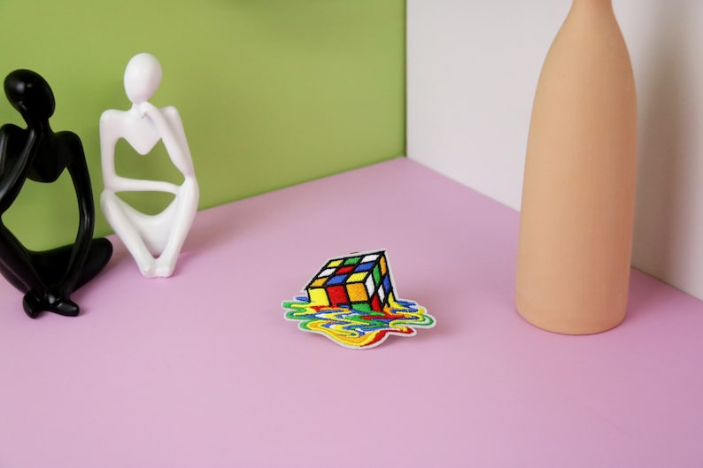 May include: A colorful embroidered iron-on patch of a melting Rubik's Cube. The patch is on a pink surface.