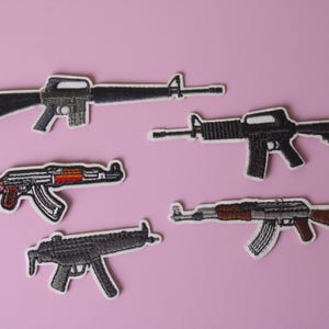 May include: Five embroidered iron-on patches of different types of rifles on a pink background.