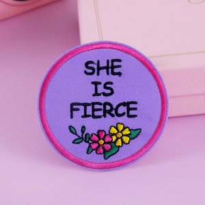 Puede incluir: Un parche redondo bordado con fondo lavanda y borde rosa intenso. El texto "SHE IS FIERCE" está en negro, sobre un diseño floral con flores rosas y amarillas.