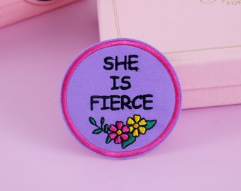 Könnte beinhalten: Ein runder, gestickter Aufnäher mit lavendelfarbenem Hintergrund und einem pinkfarbenen Rand. Der Text "SHE IS FIERCE" ist in Schwarz gehalten, über einem Blumendesign mit rosa und gelben Blumen.