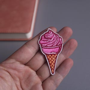 Puede incluir: Parche bordado con un cono de helado rosa. El helado es un remolino rosa, y el cono es de color naranja claro. El parche tiene un borde blanco.