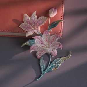Op de afbeelding: Een geborduurde patch met twee bloeiende roze leliebloemen, een gesloten lelieknop en verschillende groene bladeren. Het borduurwerk gebruikt roze, witte en blauwgroene draden om een gedetailleerd bloemmotief met zichtbare textuur te creëren.
