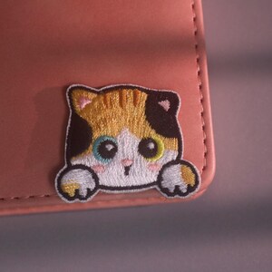 May include: An embroidered patch featuring a calico cat with large eyes, a small nose, and pink cheeks. The cat has white, orange, and black fur, and its front paws are visible as if it is peeking over the edge of a smooth, dusty pink surface.