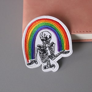 Op de afbeelding: Witte geborduurde patch met een skelet dat een regenboog vasthoudt. De regenboog is gemaakt van rood, oranje, geel, groen, blauw, indigo en violet garen.