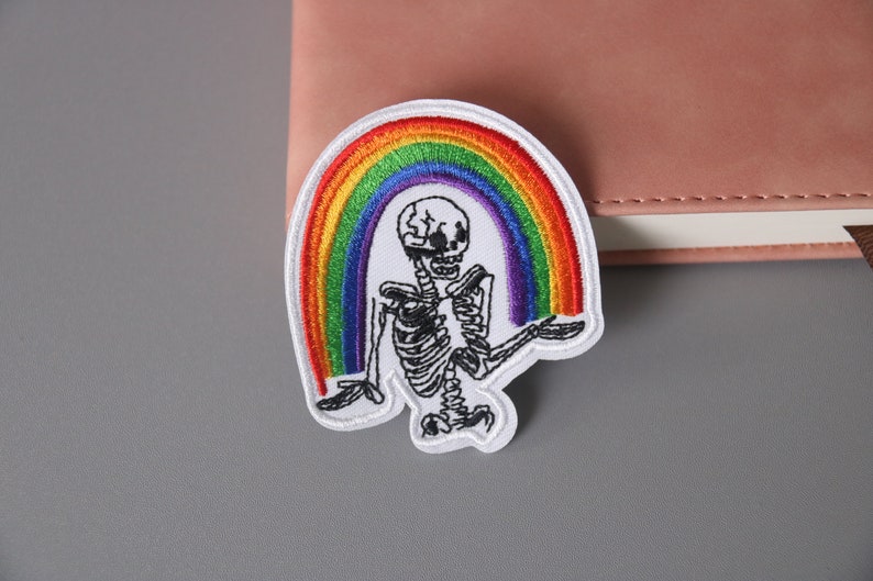 以下が含まれることがあります： 虹を持った骸骨が描かれた白い刺繍パッチ。虹は赤、オレンジ、黄、緑、青、藍、紫の糸でできています。