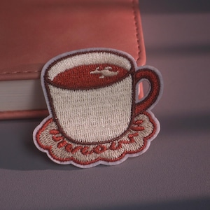 Könnte beinhalten: Ein gestickter Aufnäher mit einer cremefarbenen Teetasse, die mit dunkelroter Flüssigkeit gefüllt ist und auf einem gewellten, hellbraunen Untersetzer im Spitzenstil mit dunkelrotem Rand steht. Das Design umfasst einen dunkelroten Henkel.