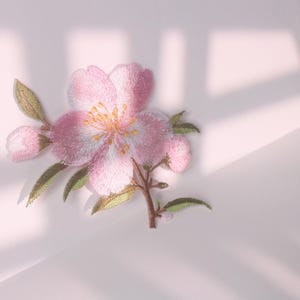 Könnte beinhalten: Gestickter rosa Blumenaufnäher mit detailliertem Design. Die Blume hat einen hellrosa Farbton mit gelbem Zentrum und grünen Blättern. Der Aufnäher dient wahrscheinlich dekorativen Zwecken, wie Kleidung oder Accessoires.