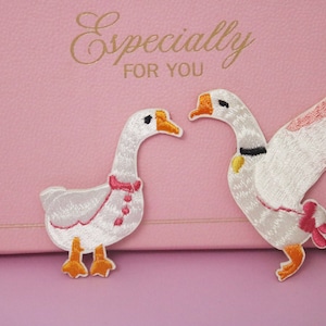 Puede incluir: Dos parches bordados de gansos. Uno tiene un lazo rosa y el otro un collar negro y un corazón amarillo. Los gansos son blancos con picos y patas naranjas. El fondo es rosa con el texto "Especially FOR YOU" en dorado.