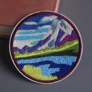 May include: Embroidered patch featuring a mountain landscape with a lake and sky. The design includes a mountain range in white, purple, and blue, a blue lake, and a green and yellow grassy area. The patch is round with a white border.
