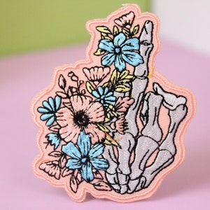 May include: A pink embroidered patch featuring a skeleton hand holding a bouquet of pink, blue, and yellow flowers.