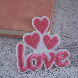 May include: White felt patch with the word "love" embroidered in pink thread. Three pink felt hearts are above the word.