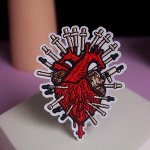 Puede incluir: Parche bordado que representa un corazón anatómico rojo atravesado por numerosas agujas plateadas y negras. El corazón tiene sombreado marrón y un borde blanco, sobre un fondo morado y blanco.