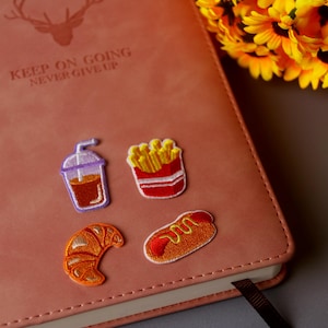 May include: A brown leather journal with the text "Keep on going never give up" embossed on the front cover.  Four embroidered patches are attached to the front cover: a cup of coffee, a carton of french fries, a hot dog, and a croissant.
