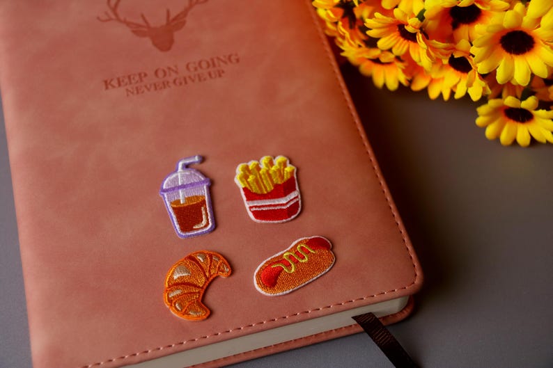 May include: A brown leather journal with the text "Keep on going never give up" embossed on the front cover.  Four embroidered patches are attached to the front cover: a cup of coffee, a carton of french fries, a hot dog, and a croissant.