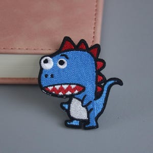 Puede incluir: Parche de dinosaurio azul bordado con púas rojas y dientes blancos. El dinosaurio está de pie sobre una superficie gris claro.