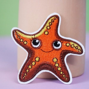 Puede incluir: Un parche bordado de estrella de mar de color marrón y naranja con detalles amarillos y grandes ojos negros. La estrella de mar sonríe y tiene un contorno blanco.