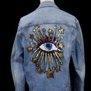 May include: A blue denim jacket with a large, colorful eye and starburst design on the back. The eye is made of sequins and has a blue iris, white sclera, and pink eyelids. The starburst is made of gold and silver sequins.
