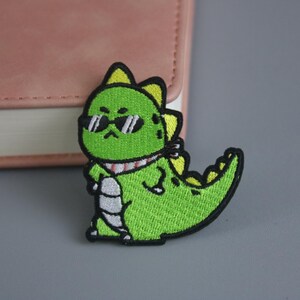 May include: Green embroidered iron-on patch of a cartoon dinosaur wearing sunglasses and a scarf.