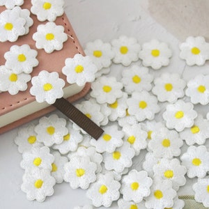 May include: A collection of white fabric daisy flower patches with yellow centres. The patches are scattered on a white surface and a pink notebook.