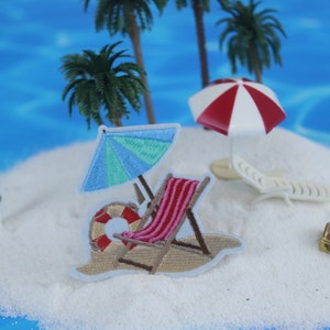 Peut inclure: Patch brodé représentant une scène de plage avec une chaise longue, un parasol et une bouée de sauvetage. Le patch est placé sur une plage de sable blanc avec des palmiers et une mer bleue en arrière-plan.