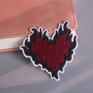 May include: Embroidered patch with a heart design in red and black, edged with white stitching. The heart is surrounded by flame-like shapes. The patch is placed on a pink leather-effect surface.