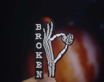 Könnte beinhalten: Weiß gestickter Patch mit einer Skeletthand, die ein gebrochenes Herz hält. Das Wort "BROKEN" ist vertikal auf schwarzem Hintergrund gestickt.