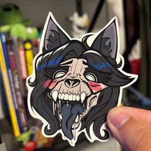 May include: A sticker featuring a stylized illustration of a wolf skull with long black hair and a blue tongue. The wolf skull has visible teeth and pink blush marks. The sticker is held by a hand.