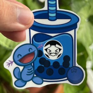 Puede incluir: Una pegatina azul con una taza de té de burbujas de dibujos animados, una criatura azul sonriente y un arándano. La taza de té de burbujas tiene una pajita y un logotipo. La criatura tiene una expresión feliz.