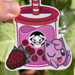Puede incluir: Una pegatina de bubble tea rosa y roja con una fresa de dibujos animados, un Pokemon rosa de dibujos animados y un logotipo en blanco y negro. El bubble tea tiene una tapa roja y una pajita blanca y roja.