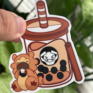 Puede incluir: Un personaje de dibujos animados marrón y blanco sosteniendo un té de boba marrón con una pajita marrón y blanca a rayas. El té de boba tiene perlas de boba negras y una cara de dibujos animados blanca con una sonrisa.