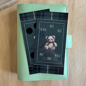 May include: A light green leather planner with two dark green and black plaid cards. One card features a teddy bear illustration and dollar amounts. The other card has black circles.