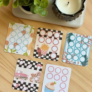 May include: Five rectangular savings challenge cards with rounded corners, each featuring a grid of empty circles for tracking progress. Designs include floral patterns, a diner theme with fries and soda, a beach scene, a retro car, and a cupcake.