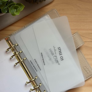Peut inclure: Agenda beige ouvert avec des anneaux dorés, contenant plusieurs inserts blancs. Les inserts affichent du texte, dont "STYLE 03", "HAIR CARE" et "PURELY BUDGETS". L'agenda est ouvert, révélant les pages intérieures.