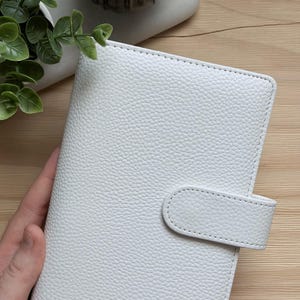 May include: A white, textured, leather-like planner with a snap closure. The planner is closed and has a stitched border. The background includes a candle and greenery.