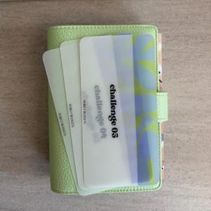May include: A light green planner with a textured cover, holding several translucent cards. The cards display the text "Challenge 04" and "Challenge 05". The planner is closed with a snap closure.