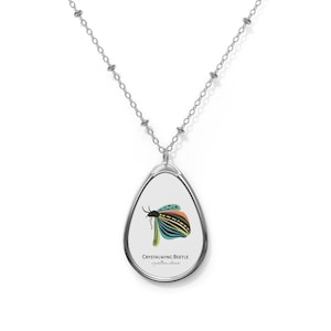 Op de afbeelding: Een zilveren ketting met een traanvormige hanger met een kleurrijke illustratie van een kever met de tekst "CRYSTALWING BEETLE" en "crystallina alatum" eronder.