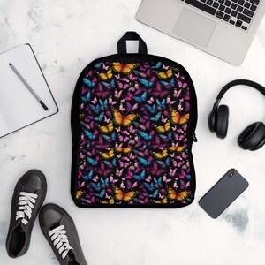 May include: Black backpack with a colorful butterfly pattern. The butterflies are in shades of pink, blue, yellow, and purple. The backpack has a zipper closure and adjustable straps.