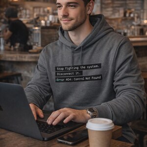 May include: A grey hoodie with the text "Stop fighting the system. Disconnect it. Error 404: Control Not Found." A person is wearing the hoodie and using a laptop. A watch and a coffee cup are on the table.