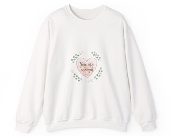 May include: A white sweatshirt with a crew neck. The front features a pink heart with the words "You are enough" in a handwritten font, surrounded by a wreath of green leaves.