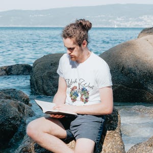 May include: A person wearing a white t-shirt with a colorful floral design of lungs and the text "Yah Weh" sitting on a rock by the ocean, reading a book.