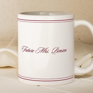 May include: A white ceramic mug featuring a maroon script text that reads Future Mrs. Bensen, accented by two thin horizontal maroon lines encircling the top and bottom of the mug, with a curved handle on the right side of the cylindrical body.