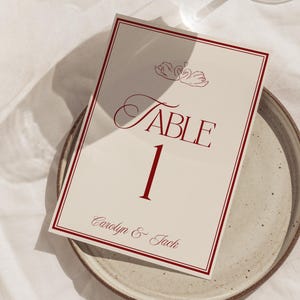 Puede incluir: Una tarjeta de número de mesa color crema con borde y texto burdeos. La tarjeta dice "Table 1" en una fuente decorativa, con los nombres "Carolyn & Jack" debajo. Dos ilustraciones de cisnes están en la parte superior. La tarjeta está sobre un plato de cerámica.