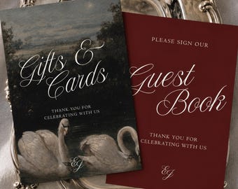 May include: Two rectangular signs resting on an ornate silver tray. The left sign features a dark landscape with two swans and the text "Gifts & Cards" and "THANK YOU FOR CELEBRATING WITH US". The right sign is deep red with "PLEASE SIGN OUR Guest Book" and "THANK YOU FOR CELEBRATING WITH US" in white script.