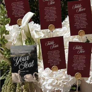 May include: Five rectangular burgundy table seating cards with white script text and gold wax seals, plus one rectangular sign featuring a swan illustration and the text "Kindly find your seat, thank you," all displayed on thin sticks.