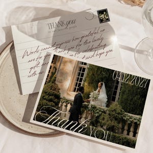 May include: A rectangular thank you card featuring a photograph of a couple in wedding attire standing on a stone balcony. The card displays the text "THANK you to our favourite people" and the date "04.12.2027" in elegant script and serif fonts.