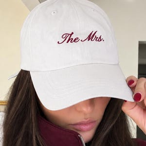 以下が含まれることがあります： 濃い赤色の筆記体で「The Mrs.」と刺繍された白い野球帽。この帽子は構造化されたクラウン、カーブしたつば、中央上部にボタンを備えています。生地は無地で不透明な素材のように見えます。