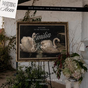 May include: A rectangular wedding sign in a gold ornate frame featuring two white swans on dark water. The text reads: WELCOME TO THE WEDDING OF EMILIA & JOVAN JULY 19TH, 2027. The sign is displayed on a black metal easel stand with floral arrangements.
