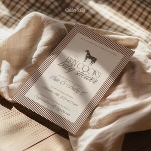 Puede incluir: Una invitaci&oacute;n a un baby shower con fondo beige y borde a rayas marr&oacute;n. La invitaci&oacute;n presenta el texto "Erin Cooks baby shower" con una ilustraci&oacute;n de caballo. El texto adicional incluye detalles del evento e informaci&oacute;n de RSVP. La invitaci&oacute;n est&aacute; colocada sobre una tela beige.