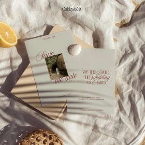 May include: Two off-white Save the Date cards with dark red serif typography and a small square photograph of a couple. The cards feature the text Oakley&Co., Save the date, November 18th, 2027, Charlotte, North Carolina, and Formal invitation to follow.