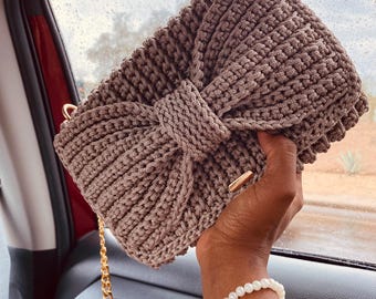 May include: A taupe-colored, crocheted handbag with a bow detail on the front. The bag has a gold chain strap and gold hardware. The handmade bag is rectangular and appears to be made of a soft yarn.
